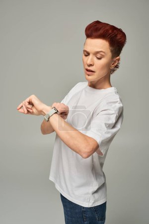 ernste rothaarige bigeschlechtliche Person im weißen T-Shirt, die die Uhrzeit auf der Armbanduhr vor grauem Hintergrund überprüft