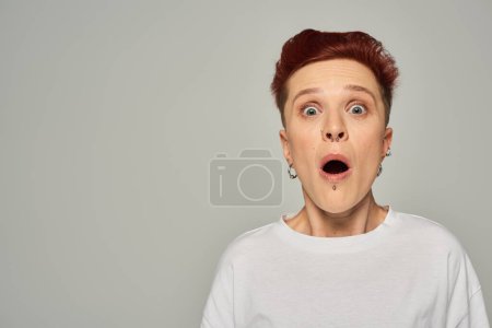 schockierte rothaarige queere Person in weißem T-Shirt mit offenem Mund und Blick in die Kamera auf grau