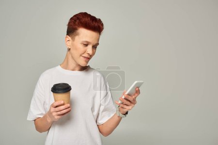 Foto de Sonriente pelirroja queer persona sosteniendo café para ir en taza de papel y mirando en el teléfono inteligente en gris - Imagen libre de derechos