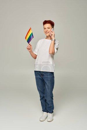 Foto de Feliz pelirroja queer persona sosteniendo pequeña bandera LGBT y hablando en el teléfono inteligente en gris, bandera - Imagen libre de derechos
