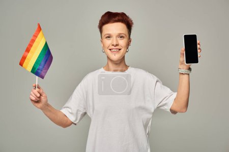 joyful redhead queer person holding small LGBT flag and smartphone with blank screen on grey