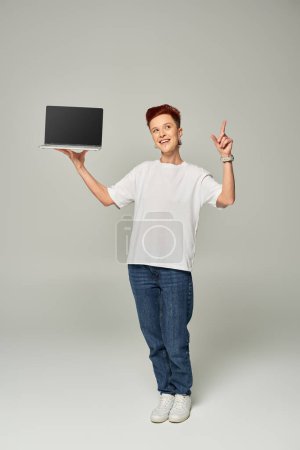 Téléchargez les photos : Sourire queer freelance montrant signe d'idée tout en tenant ordinateur portable avec écran blanc sur fond gris - en image libre de droit