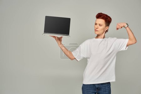 Foto de Freelancer queer confiado mostrando músculos mientras sostiene el ordenador portátil con pantalla en blanco sobre fondo gris - Imagen libre de derechos