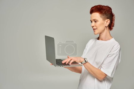 redhead queer freelancer in white t-shirt networking on laptop while standing on grey backdrop