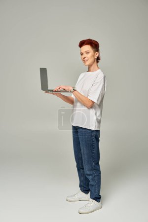 Foto de Pelirroja queer freelancer en camiseta blanca de pie con portátil y mirando a la cámara en gris - Imagen libre de derechos