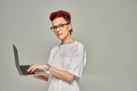 pelirroja queer freelancer en camiseta blanca y gafas de red en el portátil sobre fondo gris
