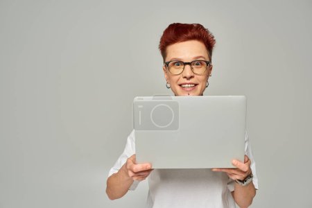 erstaunte queere Freelancerin in weißem T-Shirt und Brille, Laptop in der Hand und Kamera auf grau