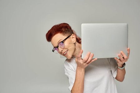 fröhliche rothaarige queere Freiberuflerin mit Brille, Laptop in der Hand und Kamera auf grau