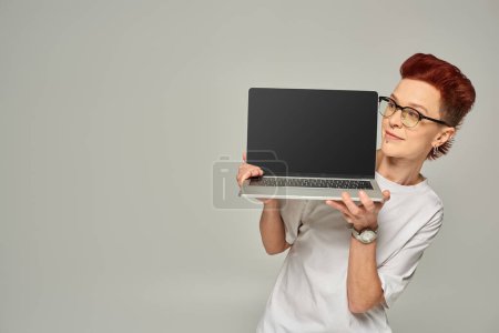 lächelnde rothaarige queere Freiberuflerin mit Brille und Laptop mit leerem Bildschirm vor grauem Hintergrund
