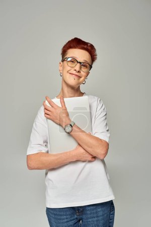 entzückte und glückliche rothaarige queere Freiberuflerin mit Brille und Laptop vor grauem Hintergrund