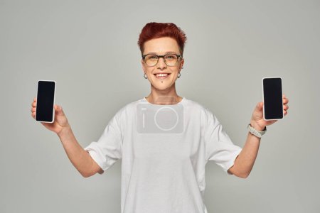 pelirroja sonriente queer persona en gafas que muestra teléfonos inteligentes con pantalla en blanco sobre fondo gris
