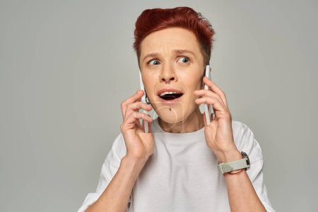 beeindruckt und erstaunt rothaarige queere Person mit offenem Mund, die auf Smartphones vor grauem Hintergrund spricht