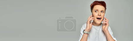 Photo for Amazed redhead queer person with open mouth talking on smartphones on grey backdrop, banner - Royalty Free Image