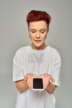 rothaarige nichtbinäre Person in weißem T-Shirt mit Handy und leerem Bildschirm auf grauem Hintergrund