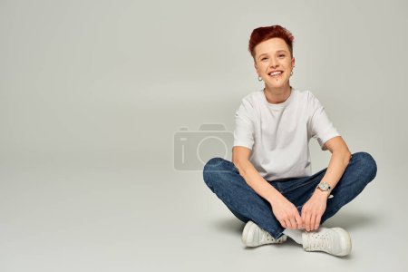cheerful redhead non-binary person in white t-shirt and jeans sitting and looking at camera on grey