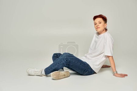 stylish redhead non-binary person in white t-shirt and jeans sitting and looking at camera on grey