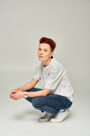 redhead queer person in white t-shirt and jeans sitting on haunches and looking at camera on grey