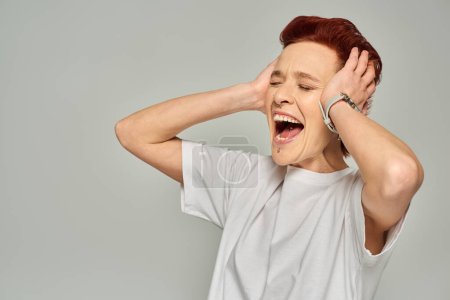 Photo for Emotional queer person in white t-shirt touching head and screaming with closed eyes on grey - Royalty Free Image