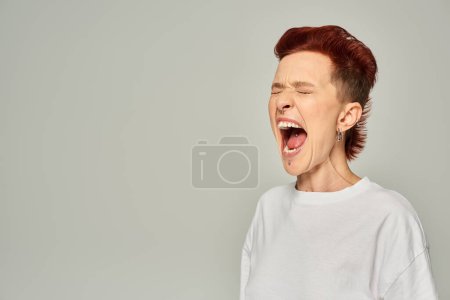 emotional queer person in white t-shirt standing and screaming with closed eyes on grey backdrop