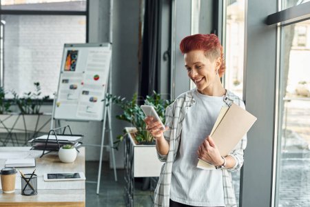 happy queer person in casual attire standing with documents in office and messaging on smartphone