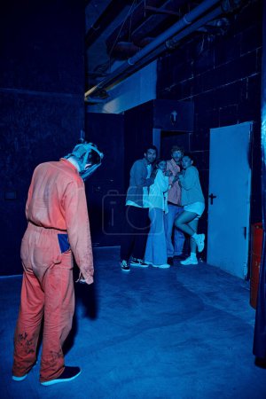 Mann in Gasmast steht in der Nähe verängstigte multikulturelle Gruppe von Menschen in Escape Room, Suche Abenteuer