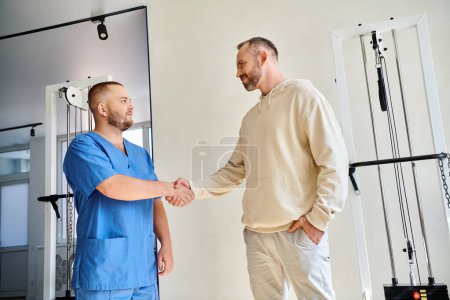 hombre sonriente estrechando la mano con el joven médico en uniforme azul en el centro kinesio rehabilitación