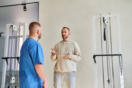 Téléchargez les photos : Patient masculin discutant du plan de traitement avec le médecin en uniforme bleu dans le centre de kinesio de récupération - en image libre de droit
