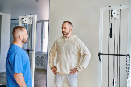 serious bearded man with hands on hips listening to young specialist in kinesiotherapy center