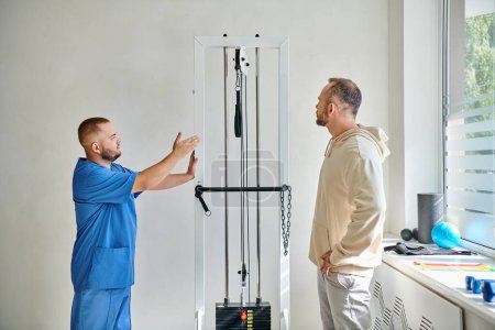 Reha-Arzt in blauer Uniform zeigt seinem Patienten im Kinesio-Zentrum Trainingsgeräte
