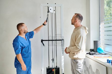 professioneller Rehabilitologe in blauer Uniform zeigt dem Mann im Kinesiologischen Zentrum Trainingsgerät