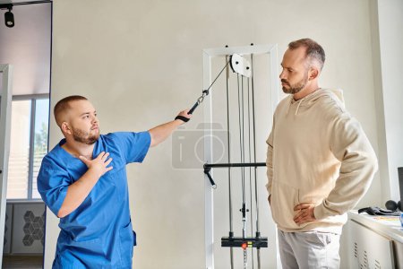 joven fisioterapeuta en uniforme azul instruyendo al hombre cerca de la máquina de ejercicio en el centro kinesio
