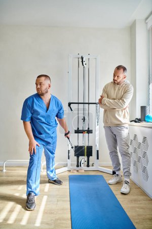 Photo for Recovery specialist in blue uniform instructing man near training machine in kinesio center - Royalty Free Image