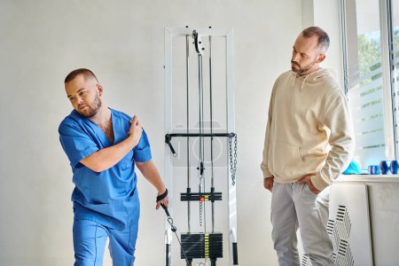 Physiotherapeut in blauer Uniform lehrt Mann in der Nähe von Trainingsgerät im Reha-Zentrum