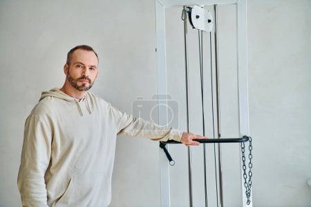 bearded handsome man looking at camera near exercise machine in gym of modern kinesiology center
