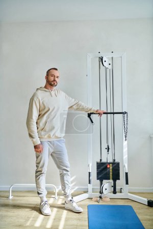 full length of handsome man near exercise machine in gym of modern kinesiology center