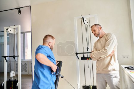 Photo for Male patient looking at physician in blue uniform training on exercise machine in kinesiology center - Royalty Free Image
