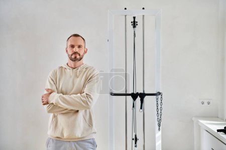 bearded man standing with folded arms and looking at camera near exercise machine in kinesio center