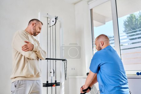 Foto de Hombre con los brazos cruzados mirando al médico joven que muestra cómo utilizar la máquina de ejercicio en el centro de recuperación - Imagen libre de derechos