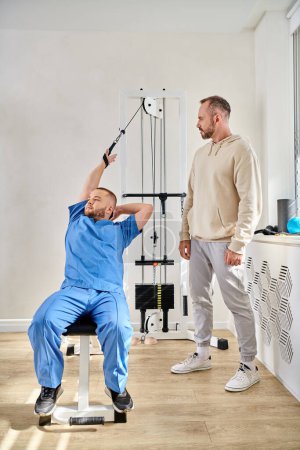 Photo for Young physiotherapist training on exercise machine during consultation of man in kinesio center - Royalty Free Image