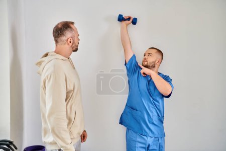 skilled rehabilitologist showing exercise with dumbbell to his patient in recovery kinesio center