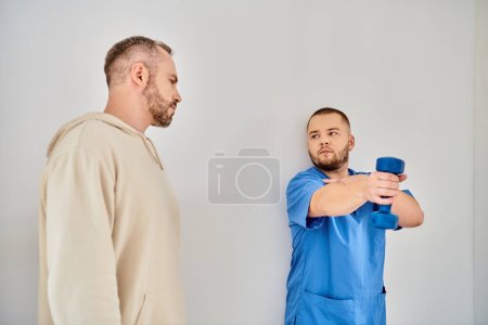 Photo for Young and skilled doctor showing how to train with dumbbell to man in recovery kinesio center - Royalty Free Image