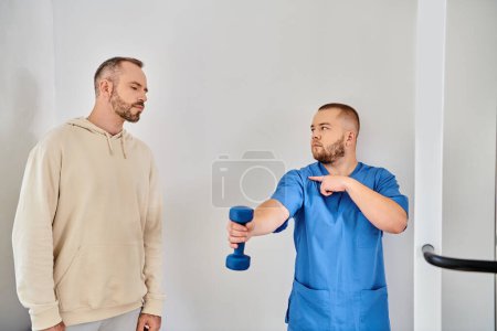 Photo for Young doctor in blue uniform showing how to train with dumbbell to man in recovery kinesio center - Royalty Free Image