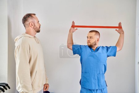 Photo for Young skilled physiotherapist showing arm exercise with resistance band to man in kinesio center - Royalty Free Image