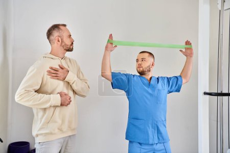 rehabilitologist showing exercise with resistance band to man with injured shoulder, kinesio center