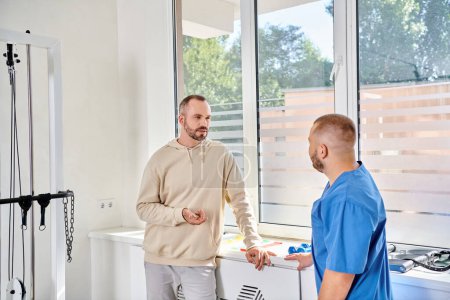 schöner Mann im Gespräch mit einem jungen Rehabilitologen in blauer Uniform während eines Termins im Kinesio-Zentrum