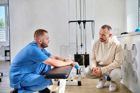 Rehabilitologe und männlicher Patient diskutieren Behandlungsplan in der Nähe von Trainingsgeräten im Kinesio-Zentrum