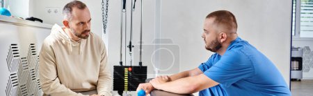 young doctor and man discussing treatment plan near exercise machine in kinesio center, banner