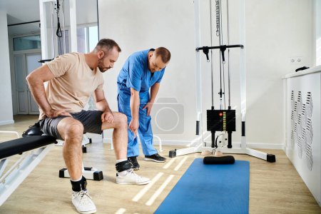 young doctor pointing at leg belts of man sitting near exercise machine in gym of kinesio center