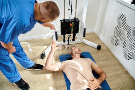 Photo for High angle view of instructor assisting his patient lying on fitness man near exercise machine - Royalty Free Image