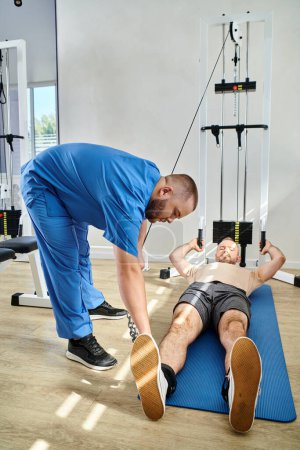 Photo for Doctor in blue uniform assisting man in sportswear lying on fitness mat near exercise machine - Royalty Free Image